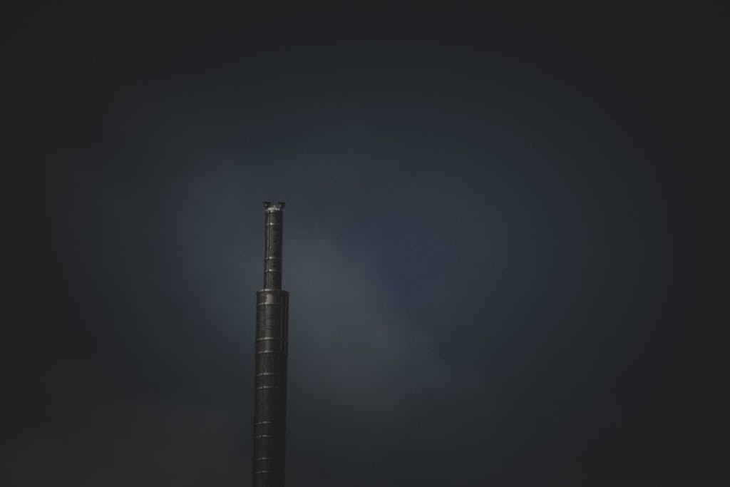 Kongotech Org A lone industrial chimney emitting smoke against a dark sky, symbolizing air pollution.