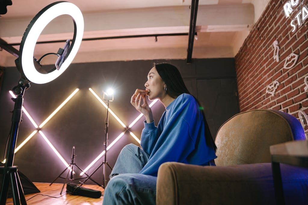 WhatsOnTech Asian woman content creator eating pizza while recording a video indoors with ring light.