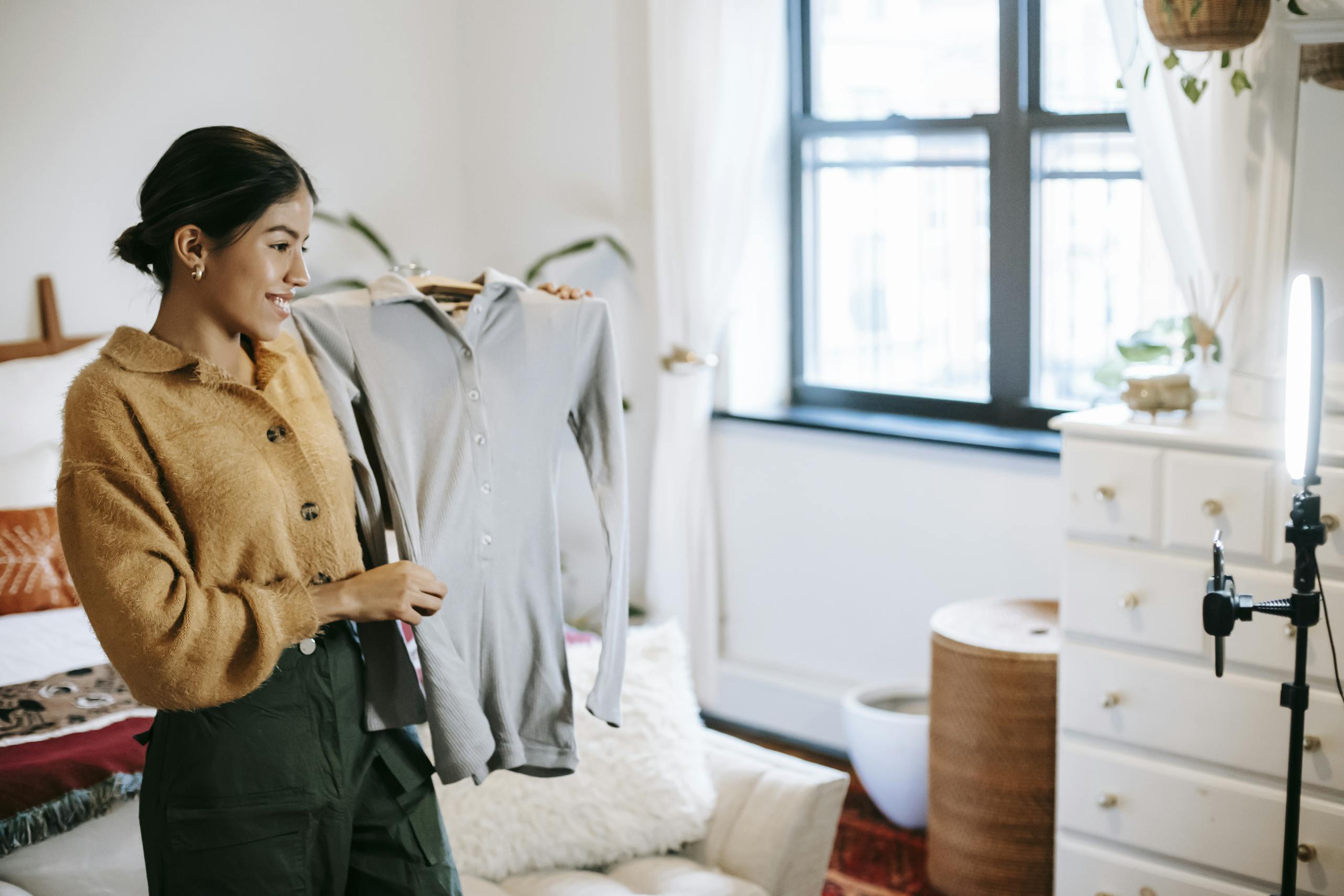 WhatsOnTech Positive ethnic female blogger demonstrating jacket on hanger during live stream on mobile phone placed on shining lamp