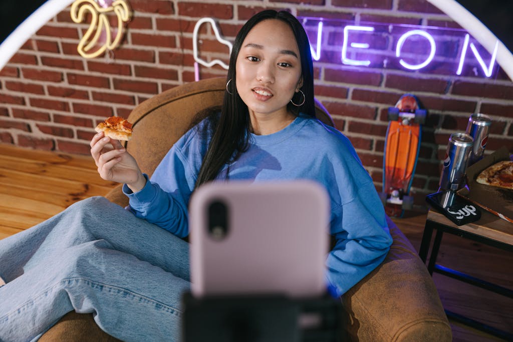WhatsOnTech Young Asian woman streaming video while enjoying pizza in a cozy setup with neon lights.