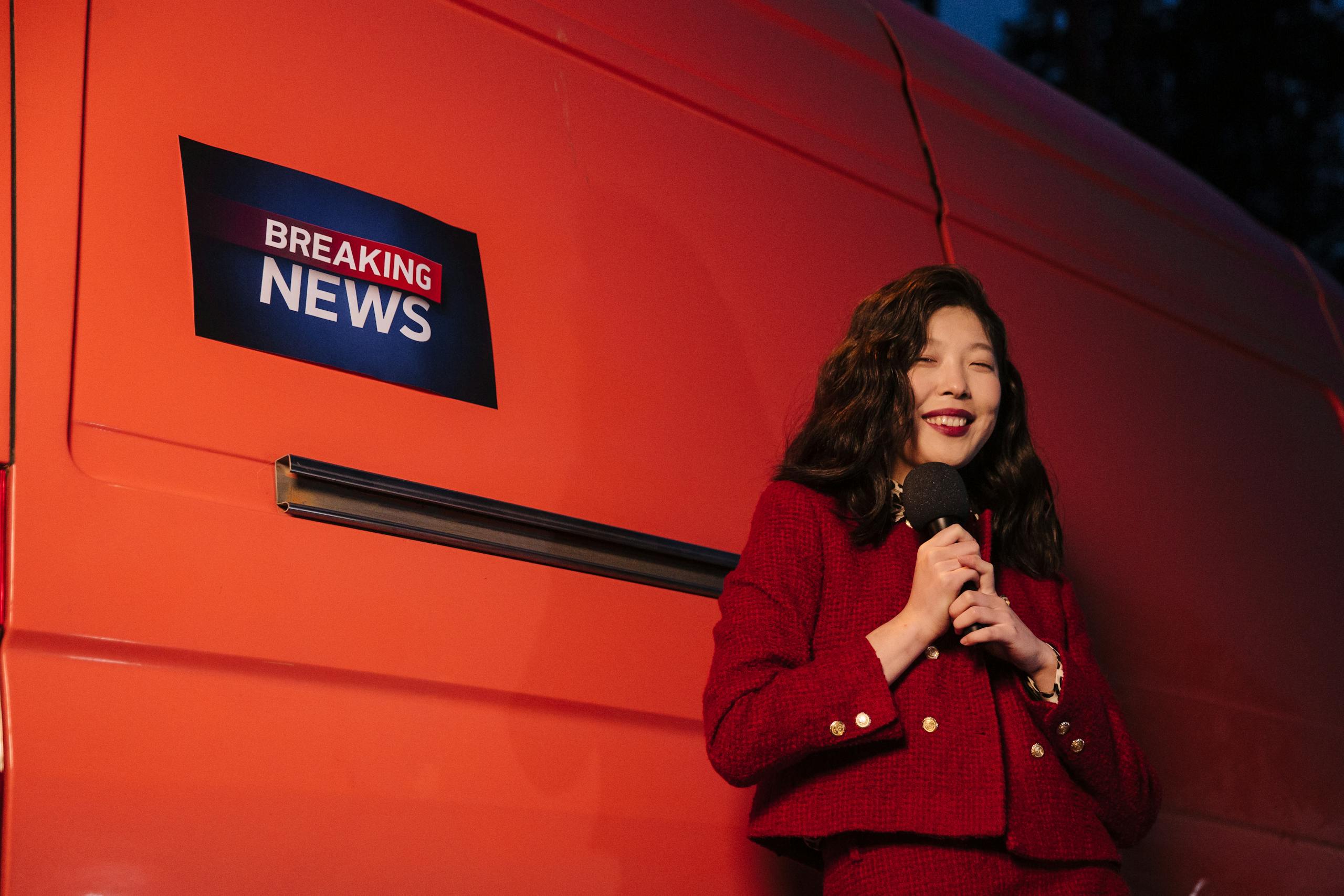 IntelligentNews.co.uk Asian female reporter smiling with microphone besides a news van at night.