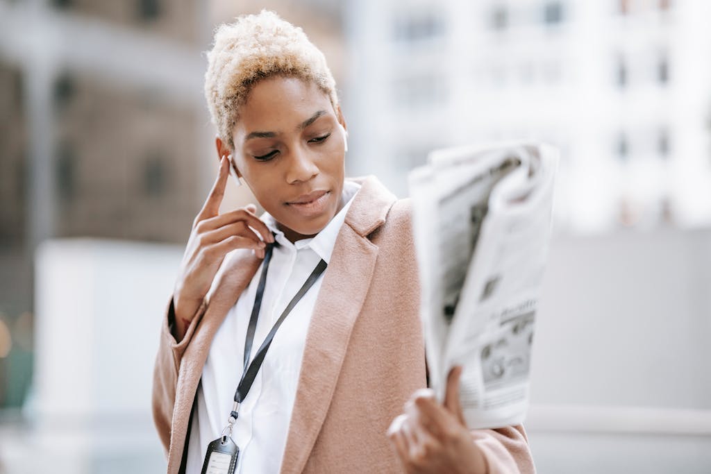 IntelligentNews.co.uk Focused young African American female entrepreneur with badge having phone call via earbuds while reading newspaper