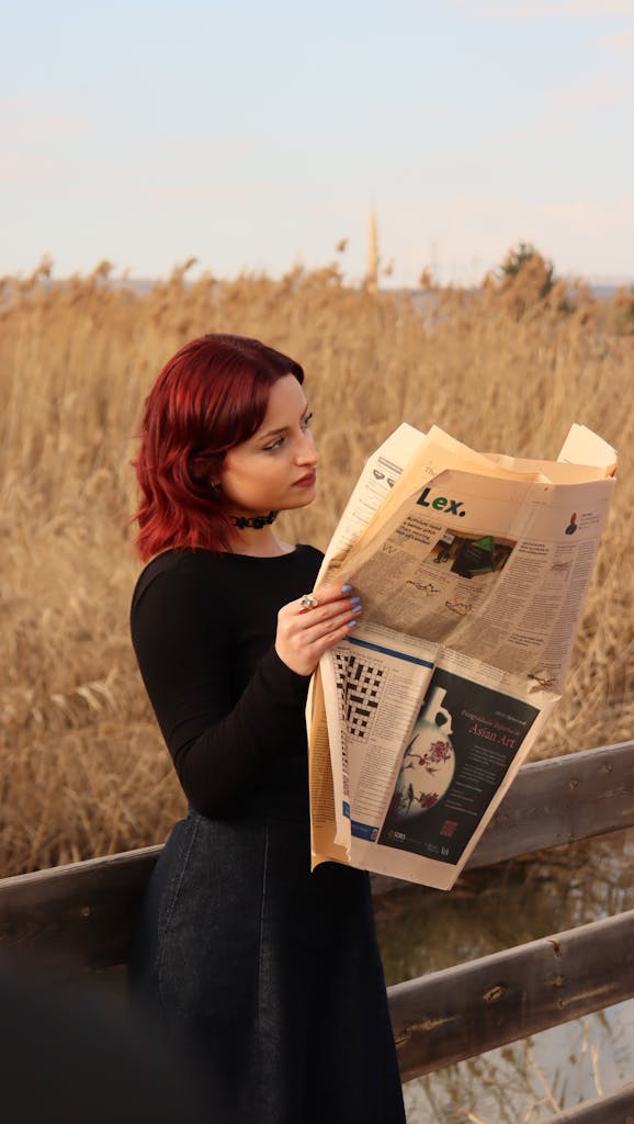 Uitly Red-haired woman reads a newspaper in a serene outdoor setting with dry reeds.