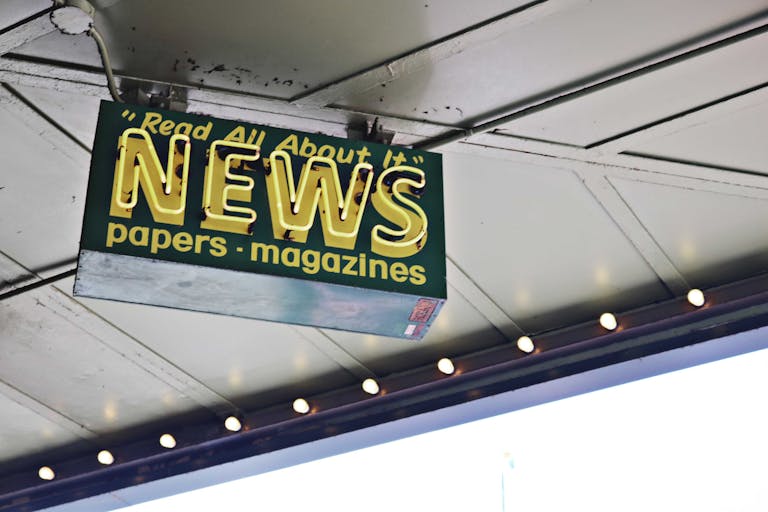 Newsatrack From below of illuminated signboard with news papers magazines inscriptions hanging on metal ceiling on street
