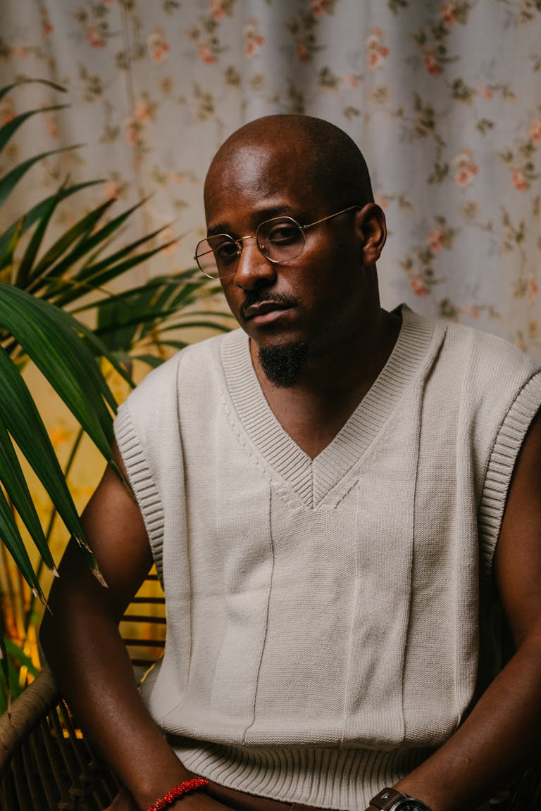 Berniece Julien Portrait of a fashionable man wearing a sleeveless sweater, sitting indoors with a potted plant backdrop.