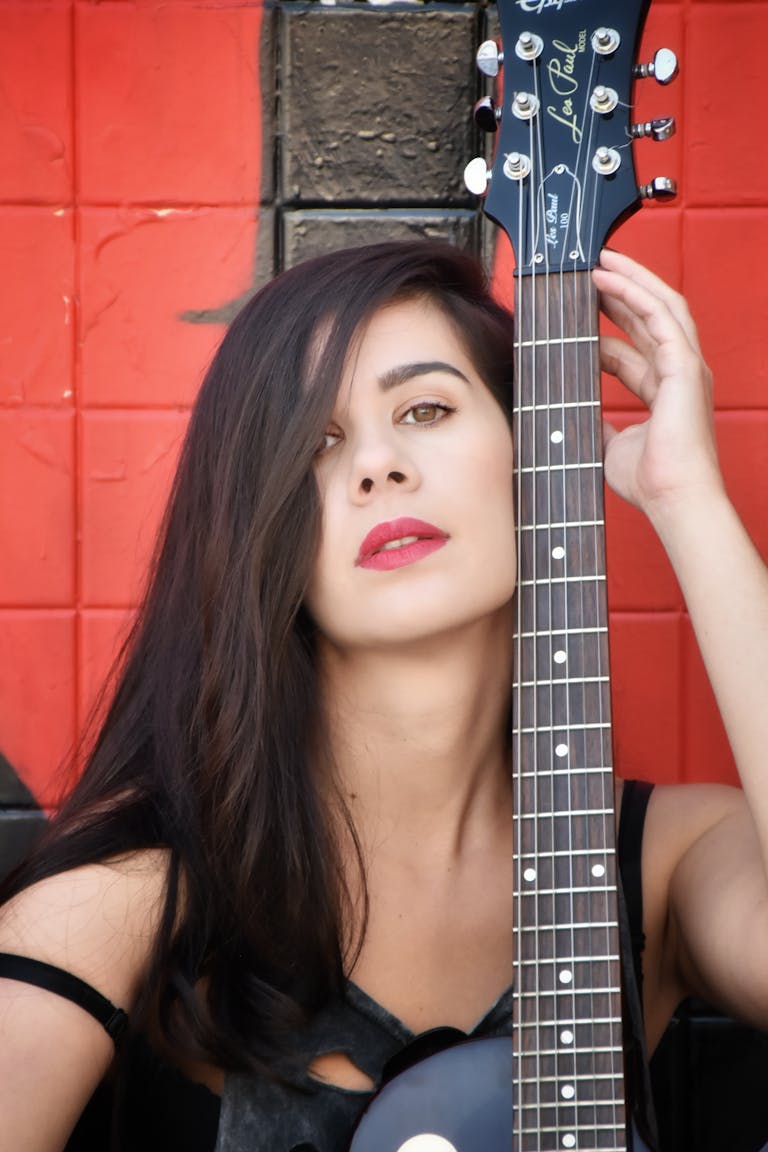 Lupe Gidley Stylish woman holding a guitar against a red backdrop, exuding modern artistry.